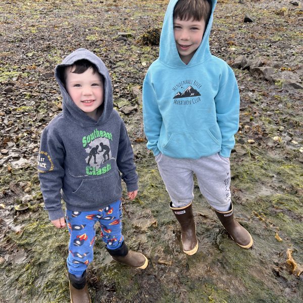 Two kids in hoodies and boots stand on a rocky, grassy shore.