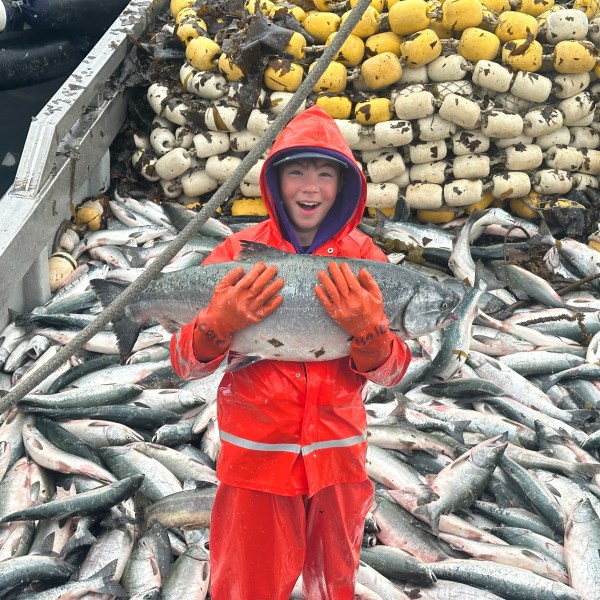 Child in orange rain gear holding fish on boat full of fish, with nets in background.
